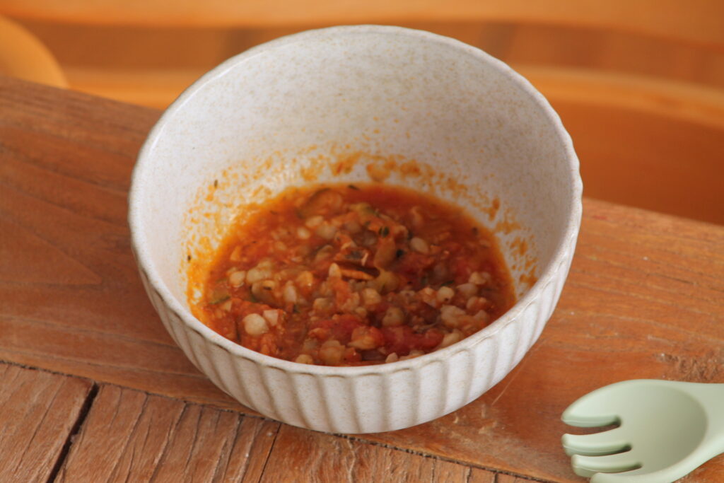 Bowl of baby food buckwheat puttanesca on table in front of highchair with baby spoon next to it